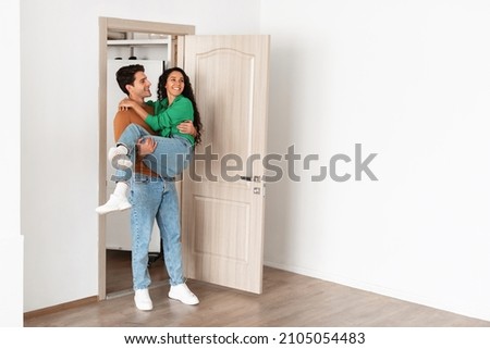 Loving millennial husband carrying wife in his arms standing in doorway, newlyweds celebrating moving day, happy family having fun in empty room, couple excited by relocation into first new house Royalty-Free Stock Photo #2105054483