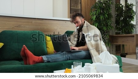 Sick male freelancer sitting on sofa at home office under plaid, working remotely, using paper tissue with termometr in mouth. Man feeling unwell, sick with running nose, need a rest. Royalty-Free Stock Photo #2105069834