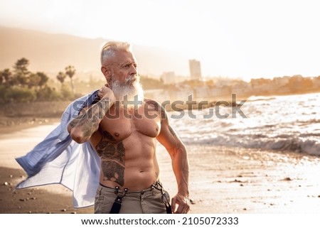 In a healthy body, healthy mind. Senior man with white stylish beard and tattoos walking on the beach at sunset. Summer vacation . Elderly people healthy lifestyle on a retirement concept. Royalty-Free Stock Photo #2105072333