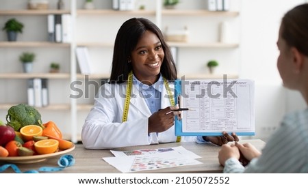 Consultation with nutritionist. Young black female dietologist showing patient example of weekly menu, having conversation at clinic. Dietitian lady showing female patient how to use diet diary Royalty-Free Stock Photo #2105072552