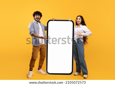 Cool mobile offer. Happy indian couple pointing at big cellphone with white screen for mockup, demonstrating free space for phone application or website design, yellow background Royalty-Free Stock Photo #2105072573