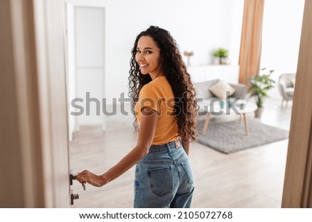 Portrait of excited young woman walking in her apartment, entering new home and looking back at camera, happy curly lady standing in doorway of modern flat, coming inside, selective focus Royalty-Free Stock Photo #2105072768