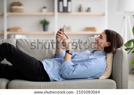 Man enjoying spare time alone lying on sofa and watching videos on cellphone, relaxing at home, using tech. Millennial guy typing on smartphone, surfing in internet in living room interior Royalty-Free Stock Photo #2105083064