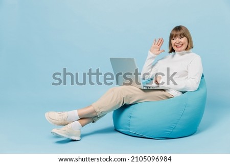 Full body elderly woman 50s in white knitted sweater sit in bag chair hold use work on laptop pc computer talk video call waving hand isolated on plain blue color background People lifestyle concept