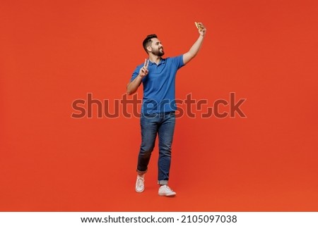 Full body young smiling happy caucasian man 20s wear basic blue t-shirt do selfie shot on mobile cell phone show v-sign isolated on plain orange background studio portrait. People lifestyle concept