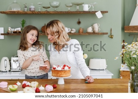 Happy chef cook baker mom woman in white shirt work with child baby girl helper use hold mobile cell phone at kitchen table home Cooking food process concept Mommy little kid daughter prepare cake.