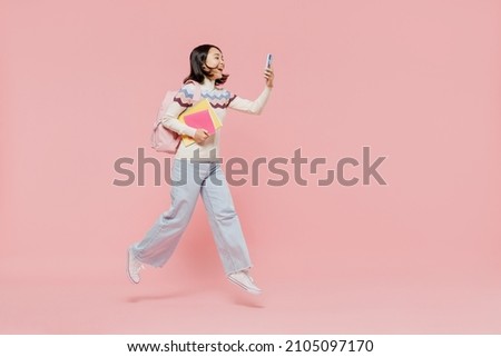 Full body teen student girl of Asian ethnicity in sweater hold backpack jump high use mobile cell phone jump high isolated on pastel plain light pink background Education in university college concept Royalty-Free Stock Photo #2105097170