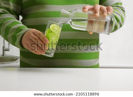 man pouring water from bottle to transparent glass  with lime slices on background  kitchen interior. no face. 