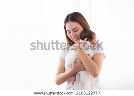 Beautiful aisan mother holding and embrace newborn baby in her arms. Woman holding adorable infant in arms and support baby back by her hand. mom and son spend time together in white room.