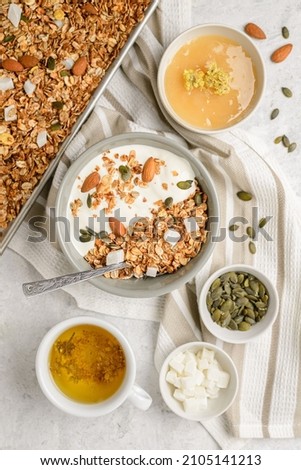 Cooked granola muesli with yogurt, honey, pumpkin seeds, almonds and coconut pieces in baking sheet and bouls with a spoon and herbal tea. Flat lay of breakfast, top view Royalty-Free Stock Photo #2105141213