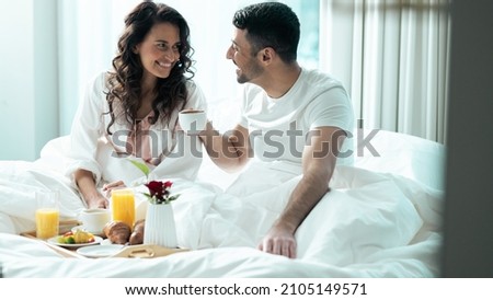Isolated Beautiful Ethnic Couple Celebrating Valentines Day in a Fancy Hotel Suite, Eating Breakfast Off of a Fancy Tray As They Stay Under The Blanket, Looking Into Each Others Eyes and Smiling. Royalty-Free Stock Photo #2105149571