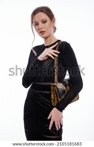 High fashion photo of a beautiful elegant young woman in a pretty long black dress with sleeve, snake skin handbag posing on white background. Studio shot. Slim figure, gathered back hair