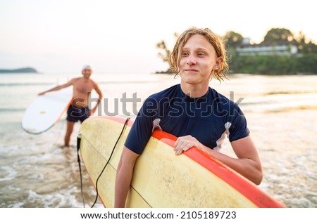 Portrait of a teen boy with a surfboard after surfing with his father. They are smiling and walking out of the water. Family active vacation concept. Royalty-Free Stock Photo #2105189723