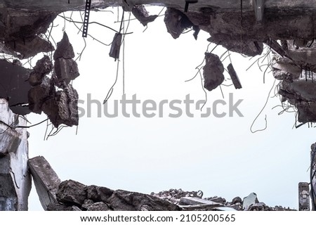 A hole in the body of a building with a pile of construction debris and concrete fragments hanging on the rebar against a uniform gray sky. Background. Royalty-Free Stock Photo #2105200565