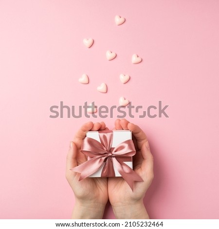 First person vertical top view photo of valentine's day decorations woman's hands holding small white giftbox with pink ribbon bow on palms and small pink hearts on isolated pastel pink background