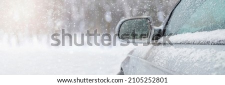 The car drives through the snow, the bright winter sun shines against the background of the forest. Close-up rearview mirror. The vehicle is covered with snow. Snowing. Royalty-Free Stock Photo #2105252801
