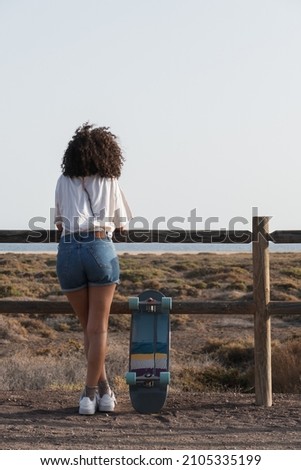 Young skater woman with afro hair from behind. She is leaning on a fence watching the sea