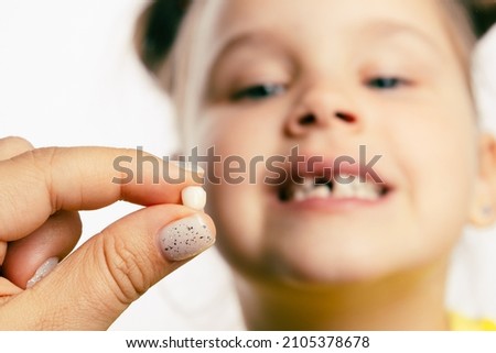 Fingers holding torn out baby milk tooth with little blurred girl face looking at tooth and showing teeth behind on white background. First teeth changing. Going to dentist to do tooth treatment.  Royalty-Free Stock Photo #2105378678