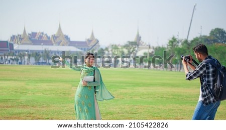 A young couple of South Asian tourists are happily posing for pictures at Sanam Luang behind the Temple of the Emerald Buddha during a long weekend. in Bangkok, Thailand