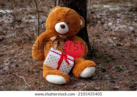 Teddy bear with gift box and heart is sitting in winter forest under tree and waiting for his couple in love. Сoncept of Valentine's Day. Romance and love. Acquaintances and confessions. Tenderness.