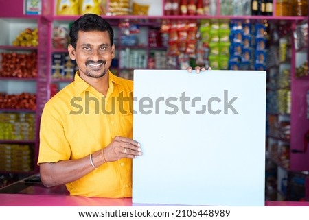 Kirana or groceries Merchant or shopkeeper with Empty sign borad looking at camera - concept of advertisement, promotions and offers on retail store Royalty-Free Stock Photo #2105448989