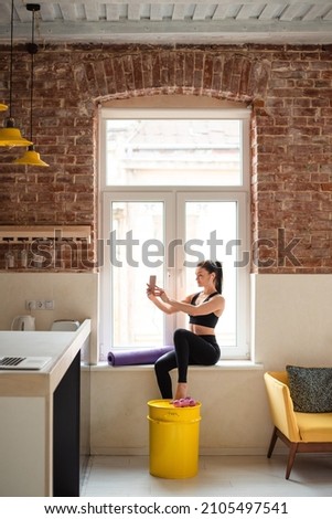 Sporty woman sitting on windowsill and using modern smartphone and earphones for listening music. Young lady in activewear resting after domestic workout.