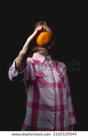 Little cute girl posing with tangerine in the studio. Photo isolated on black background with one light source. The child covers his face with tangerine.