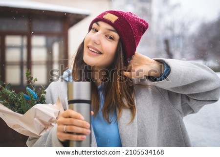 A girl with a thermos in her hands walks under the snow. Enjoy the snowfall and snowy weather. A beautiful girl smiles and enjoys the snowy weather. Snowy winter.