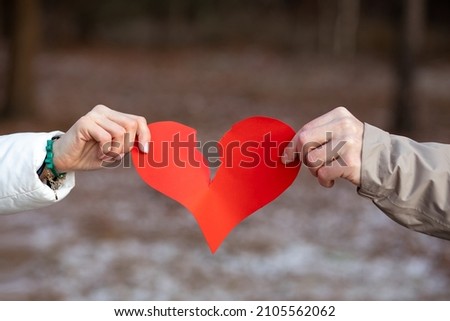 Two hands, male and female, tear red heart symbol of lovers against background of winter forest. Сoncept of breaking up relationship. Love is over. Resentment and separation. Negative. Royalty-Free Stock Photo #2105562062