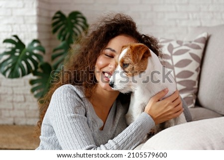 Portrait of young beautiful black woman with her adorable wire haired Jack Russel terrier puppy at home. Loving girl with rough coated pup having fun on the couch. Background, close up, copy space. Royalty-Free Stock Photo #2105579600