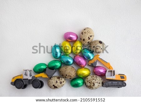 models of toy excavator and loader, lot of chocolate eggs in colorful wrappers, quail eggs on light background. concept of business congratulations on Easter holiday for construction companies.