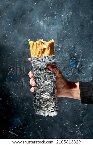 A hand holds a bitten shawarma in foil on a black textured background. Kebab with meat