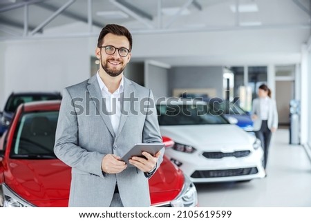 Smiling friendly car seller in suit standing in car salon and holding tablet. It's always pleasure to buy a car on a right place. Royalty-Free Stock Photo #2105619599