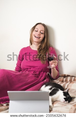 Young millennial woman in pink dress with long hair drink wine and using a tablet with her black and white tuxedo cat at home.
