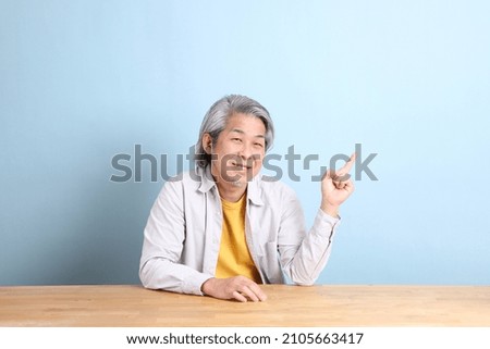 The senior Asian man with grey shirt sitting at the working desk with the blue background.