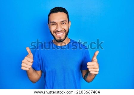Hispanic man with beard wearing casual blue t shirt success sign doing positive gesture with hand, thumbs up smiling and happy. cheerful expression and winner gesture.  Royalty-Free Stock Photo #2105667404