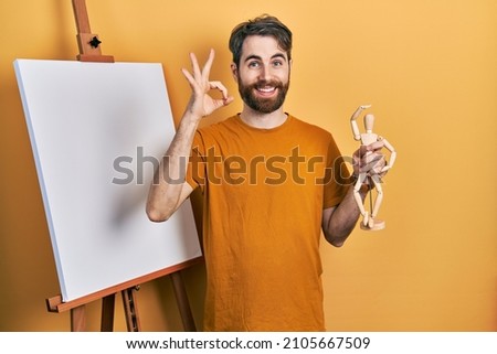 Caucasian man with beard standing by painter easel stand holding manikin doing ok sign with fingers, smiling friendly gesturing excellent symbol 