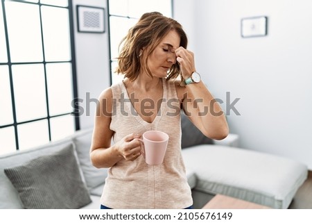 Middle age woman drinking a cup coffee at home tired rubbing nose and eyes feeling fatigue and headache. stress and frustration concept.  Royalty-Free Stock Photo #2105676149