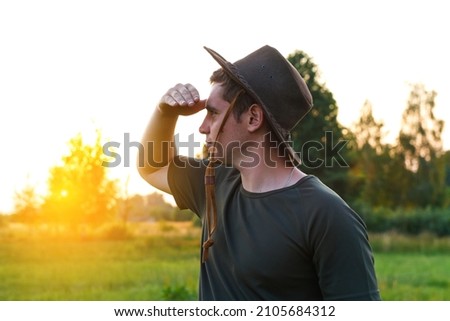 Young man farmer in cowboy hat at agricultural field on sunset with sun flare. Closeup portrait of millennial man with hat, standing on nature background, outdoors. Rancher look in to distance. 