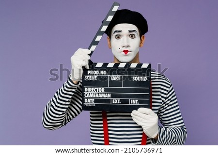 Charismatic amazing vivid young mime man with white face mask wears striped shirt beret holding classic black film making clapperboard isolated on plain pastel light violet background studio portrait