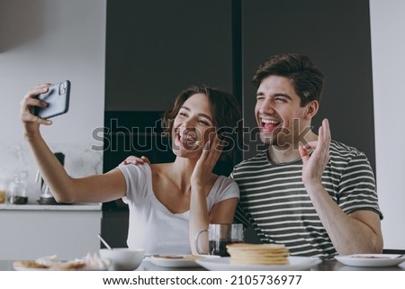 Young excited couple two woman man 20s in casual clothes sit by table eat pancake do selfie by mobile cell phone hold face show okay cook food in light kitchen at home together Healthy diet concept