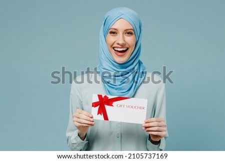 Young arabian asian muslim woman in abaya hijab hold gift certificate coupon voucher card for store isolated on plain blue background studio portrait. People uae middle eastern islam religious concept