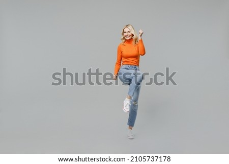 Full body elderly overjoyed excited happy blonde woman 50s in orange turtleneck do winner gesture raise up leg clench fist isolated on plain grey background studio portrait. People lifestyle concept Royalty-Free Stock Photo #2105737178