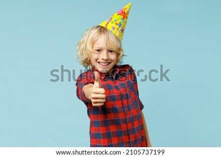 Cheerful little kid boy 10s years old in basic red checkered shirt birthday hat showing thumb up looking camera isolated on blue color background children studio portrait. Childhood lifestyle concept
