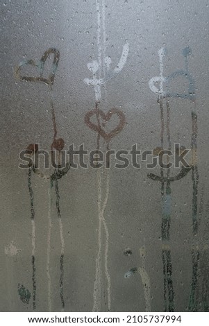 steamed glass, hand drawn smiling face and heart shapes on steamed glass surface or window with rain drops in rainy day. background of raindrops on windowpane with selective focus and noise effects