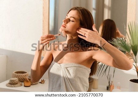 Portrait of a beautiful woman wrapped in a white towel to massage her face with a Y-shaped ball roller. Natural cosmetics concept, wrinkle smoothing Royalty-Free Stock Photo #2105770070