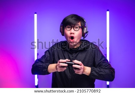 Excited and shocked face of Asian gamer with headphone playing computer pc video game online sitting at living room. Young professional gamer streaming on social playing game very fun