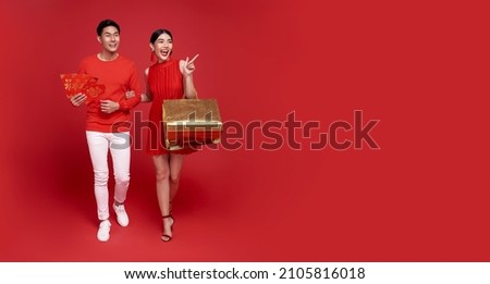 Happy Asian couple holding shopping bags and red envelopes isolated on red background for Chinese new year shopping concept. Royalty-Free Stock Photo #2105816018