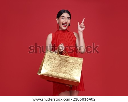 Smiling beautiful Asian woman holding shopping bags and pointing to copy space isolated on red background for Chinese new year sale concept. Royalty-Free Stock Photo #2105816402