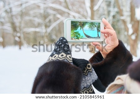 A girl in a snowy park takes pictures on a smartphone. Warm sea on the smartphone screen.